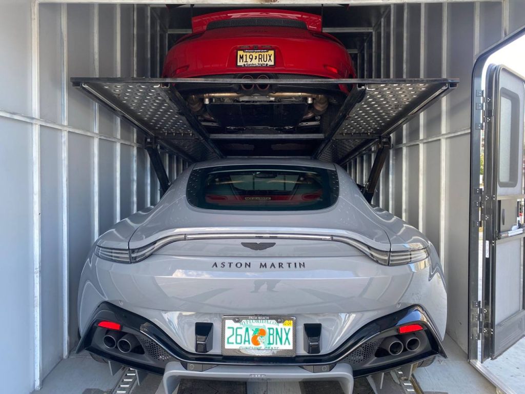 A gray Aston Martin sports car is securely positioned in the lower level of a car trailer, with a red sports car above it on a hydraulic lift.